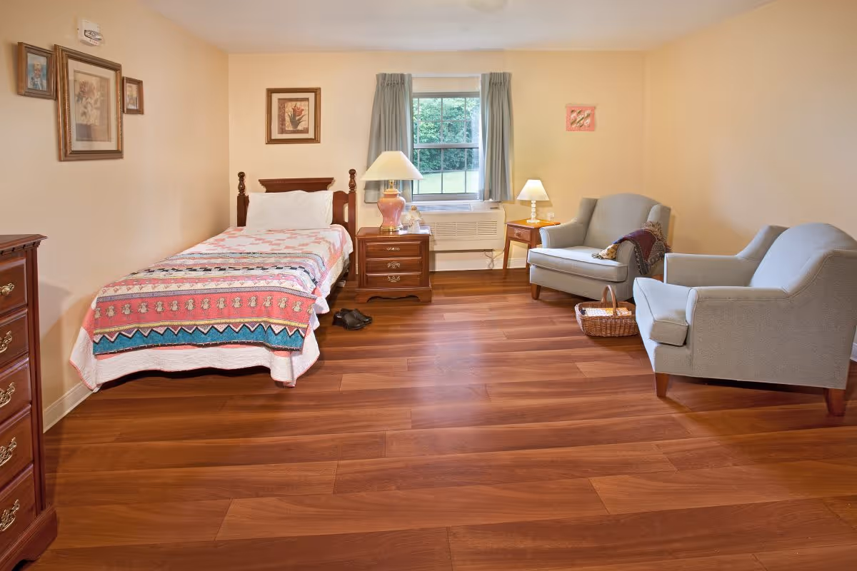 A cozy bedroom with a single bed covered in a colorful quilt, a wooden nightstand with a lamp, two gray armchairs, a small side table with a lamp, and framed artwork on the walls. There is a window with curtains letting in natural light and wooden flooring throughout the room.