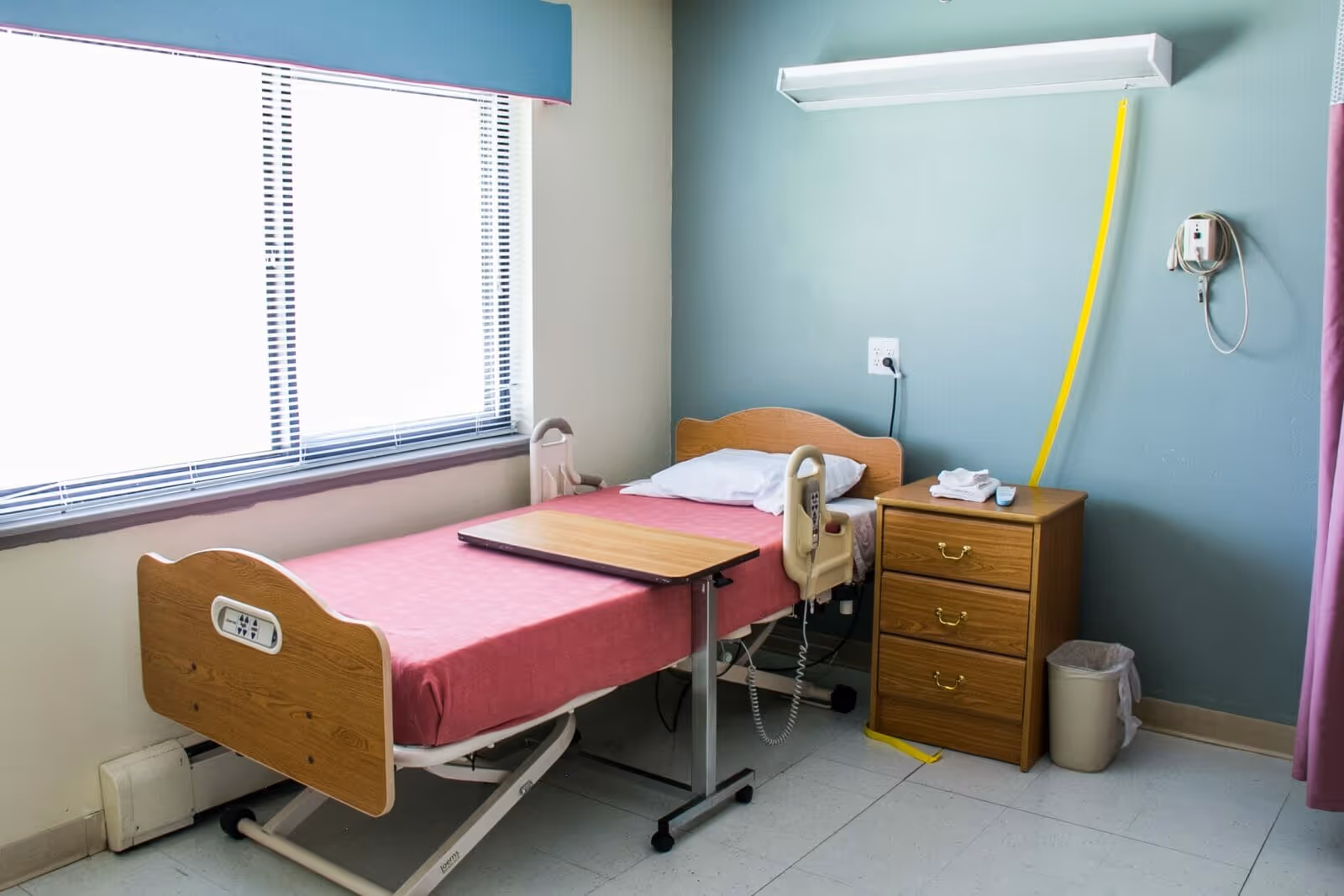 A simple patient bedroom with a single adjustable hospital bed, overbed table, wooden bedside dresser, window, and medical call device on the wall.