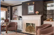 A cozy living room area featuring a white fireplace with a decorative screen, flanked by built-in white shelves filled with books and decorative items. There are three mauve upholstered armchairs arranged around the fireplace, a small side table with a lamp, and a framed picture hanging above the fireplace. The room has a warm and inviting atmosphere.