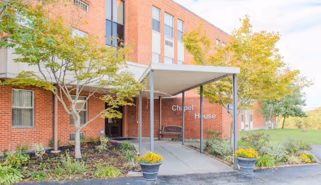 Exterior view of a brick building with a covered entrance labeled 'Chapel House'. There are trees and flower pots with yellow flowers near the entrance, and a bench under the covered area.