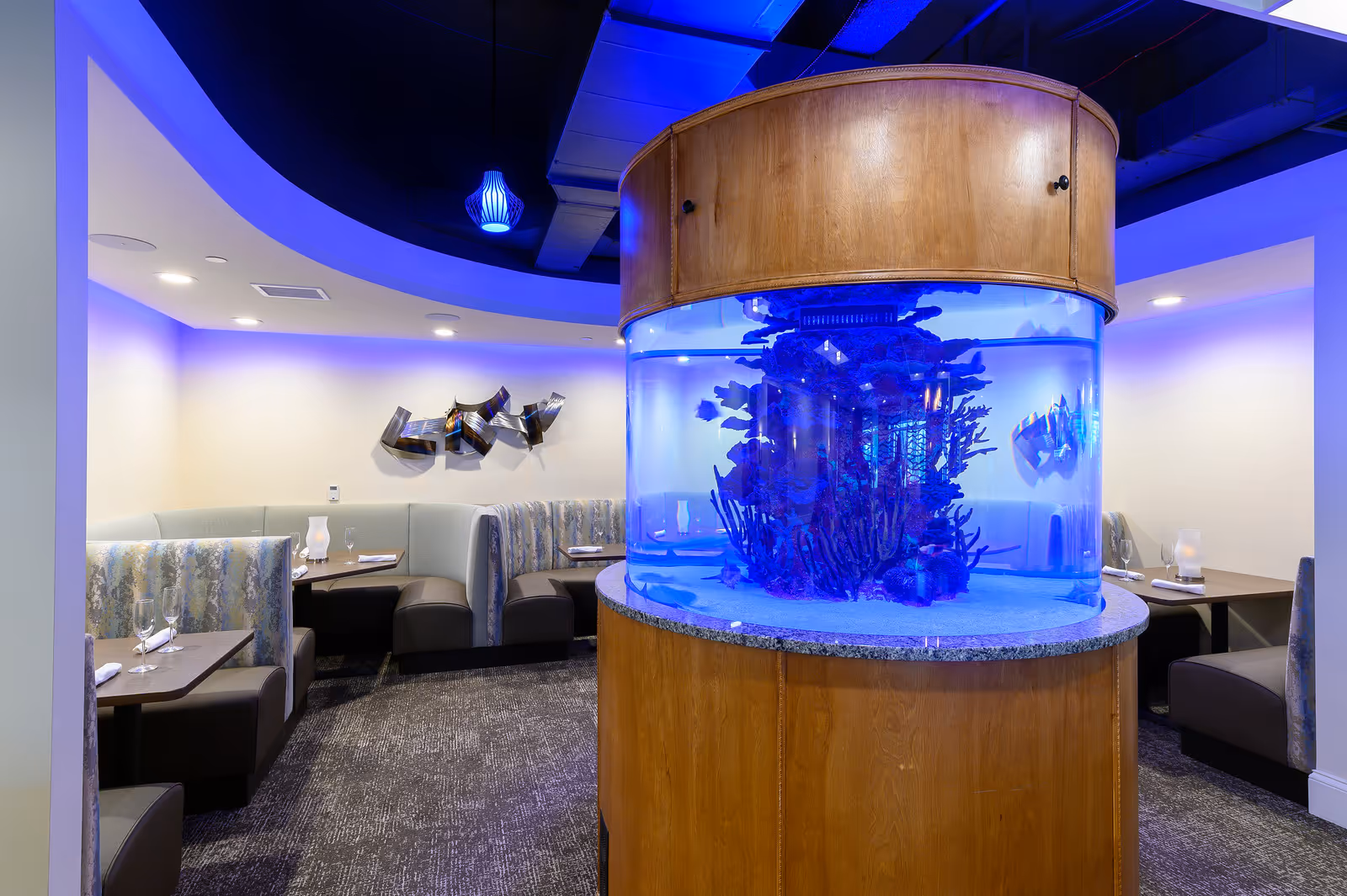 Interior view of a dining area in a senior living facility featuring curved booth seating with tables set with glasses and napkins. A large cylindrical aquarium with blue lighting and coral decorations is prominently placed in the center. The ceiling has blue accent lighting and a modern hanging light fixture.