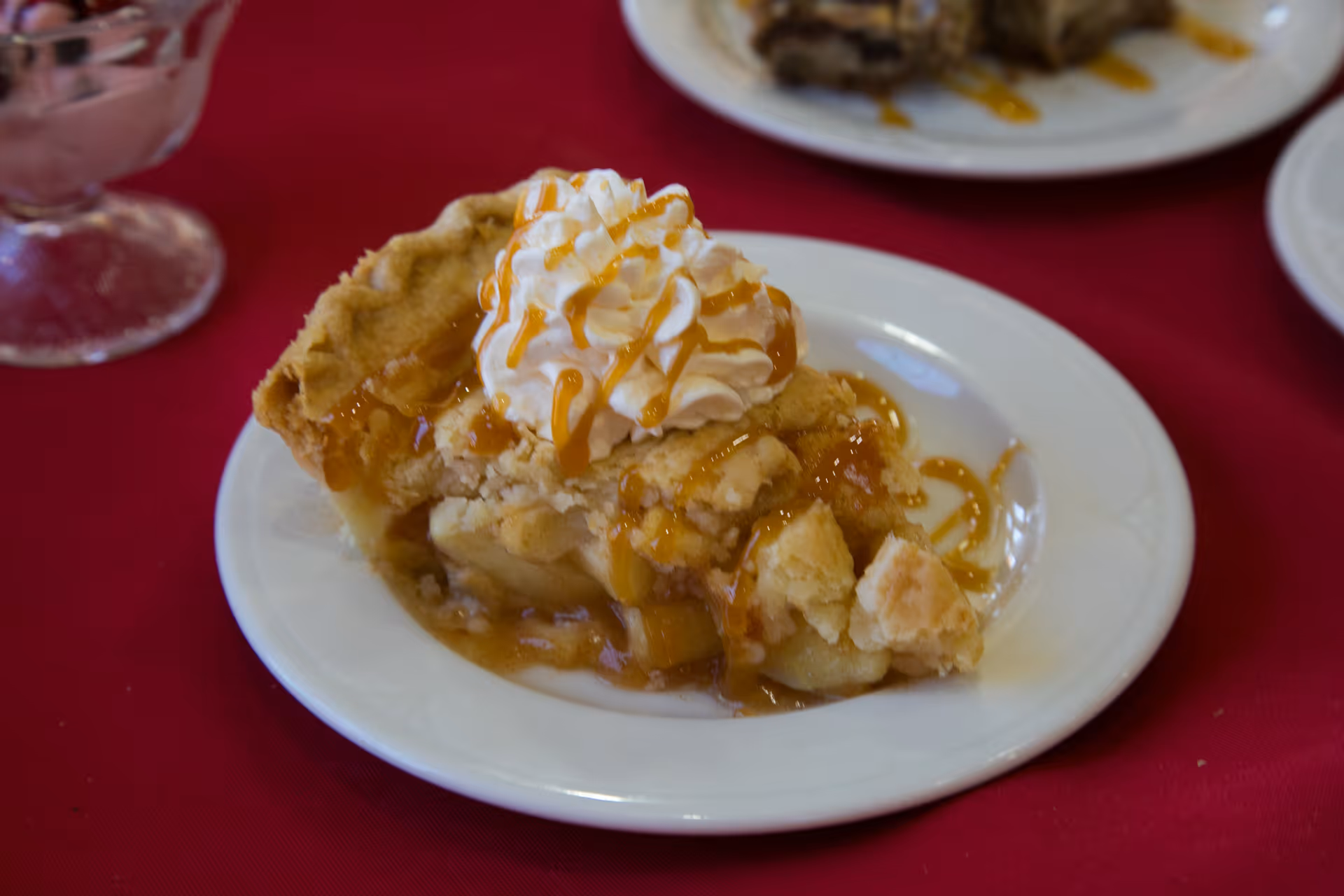 A slice of apple pie topped with whipped cream and caramel drizzle on a white plate, placed on a red tablecloth.