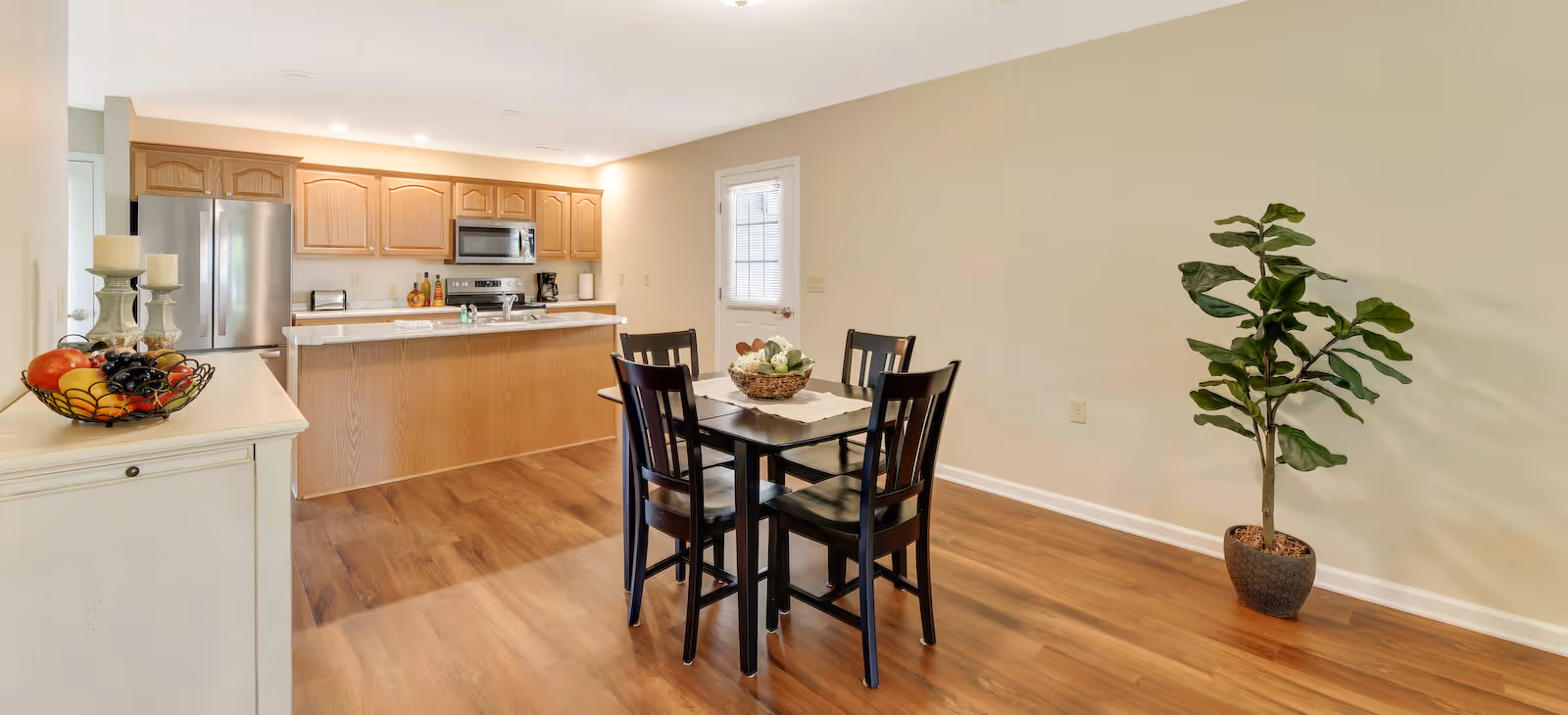 A bright kitchen and dining area with wooden flooring. The kitchen features wooden cabinets, a stainless steel refrigerator, microwave, and stove. In the dining area, there is a small square table with four black chairs and a decorative centerpiece. A potted plant is placed against the wall, and a sideboard with a fruit basket and candles is visible on the left side. A door with a window is seen in the background.