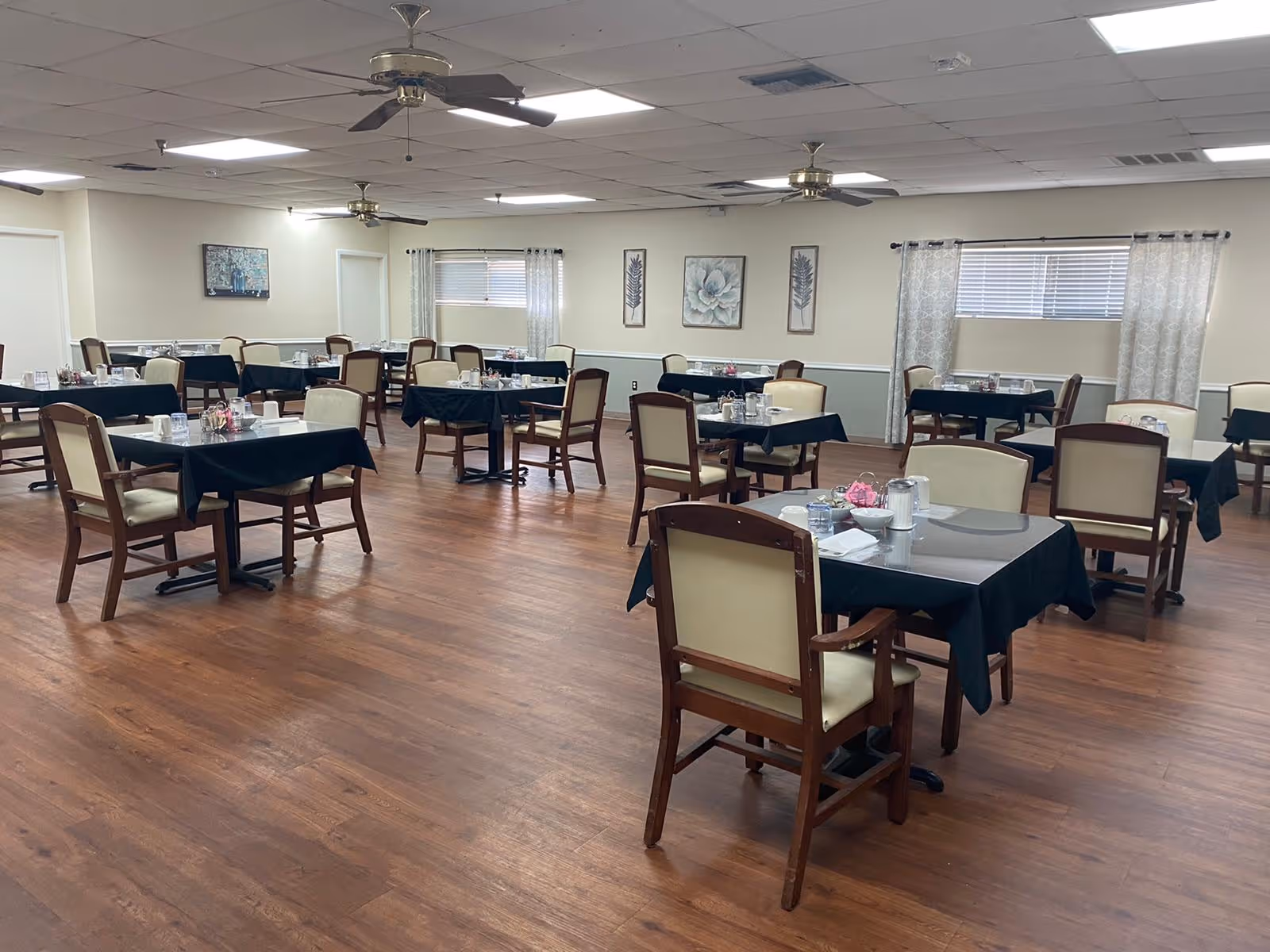 Spacious dining room with multiple tables set for meals, wooden chairs, ceiling fans, and wood flooring.