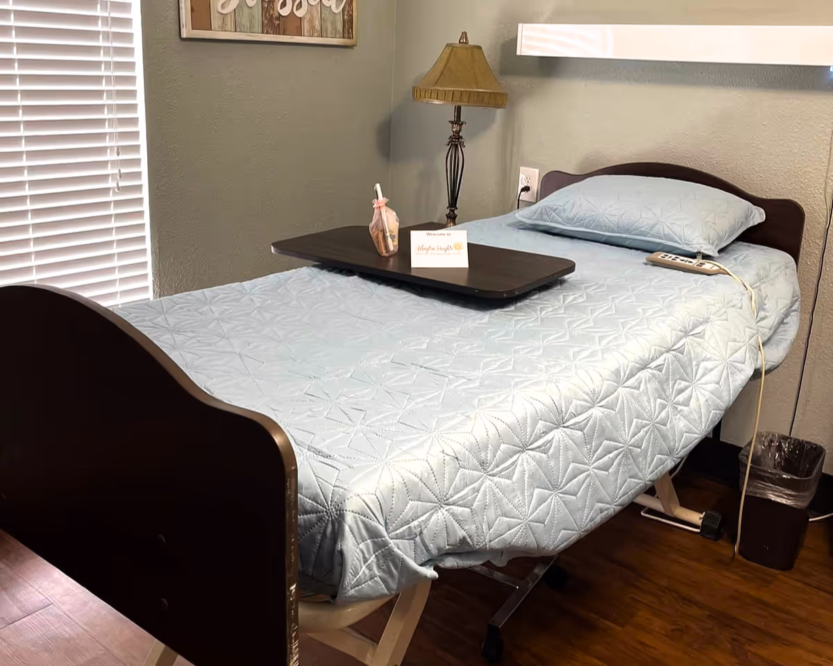 A neatly made hospital-style bed with a light blue quilted bedspread and matching pillow in a room with wooden flooring. A dark wooden overbed table is placed on the bed with a small decorative item and a welcome card. A lamp with a beige shade is on a small table next to the bed. The room has a window with white blinds and a framed wall decoration above the bed.
