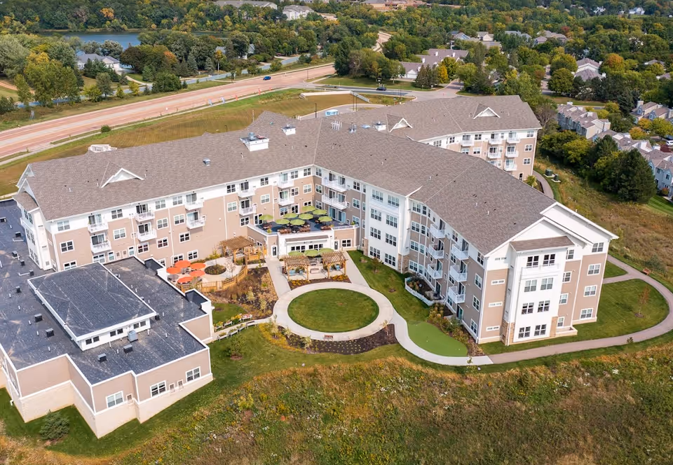 Aerial view of Riley Crossing Senior Living facility showing a large multi-story building with beige and white exterior, surrounded by landscaped green areas, walking paths, and outdoor seating with umbrellas. The building is situated near a road and residential neighborhood with trees and greenery in the background.