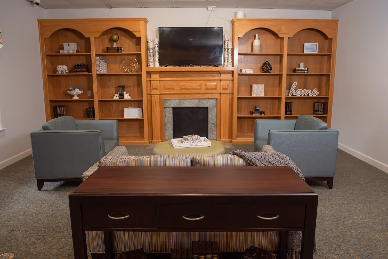 A cozy living room with two blue armchairs and a striped sofa arranged around a green ottoman. Behind the seating area is a wooden console table. The room features a wooden fireplace mantel with a flat-screen TV mounted above it, flanked by two wooden bookshelves filled with decorative items and books. The walls are light-colored and the floor is carpeted.