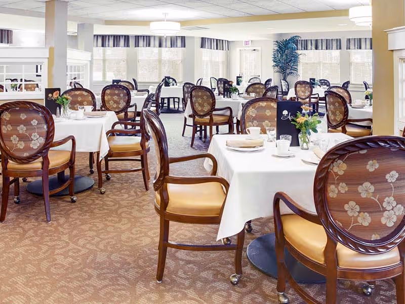 A bright and elegant dining room with multiple round tables covered with white tablecloths. Each table is set with plates, cups, and small flower arrangements. The chairs have wooden frames with floral patterned backs and tan cushions. Large windows with striped valances allow natural light to fill the room, and there are ceiling lights providing additional illumination.