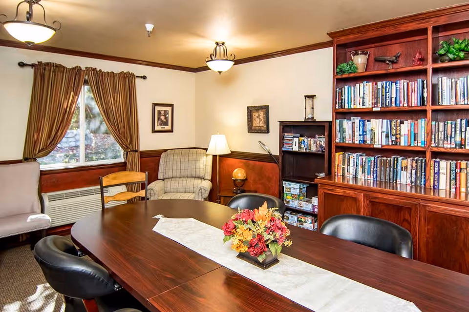 A cozy room with a large wooden table surrounded by chairs, a floral centerpiece on the table, a bookshelf filled with books and board games, a comfortable armchair, framed pictures on the wall, a window with brown curtains, and warm lighting from ceiling fixtures and a floor lamp.