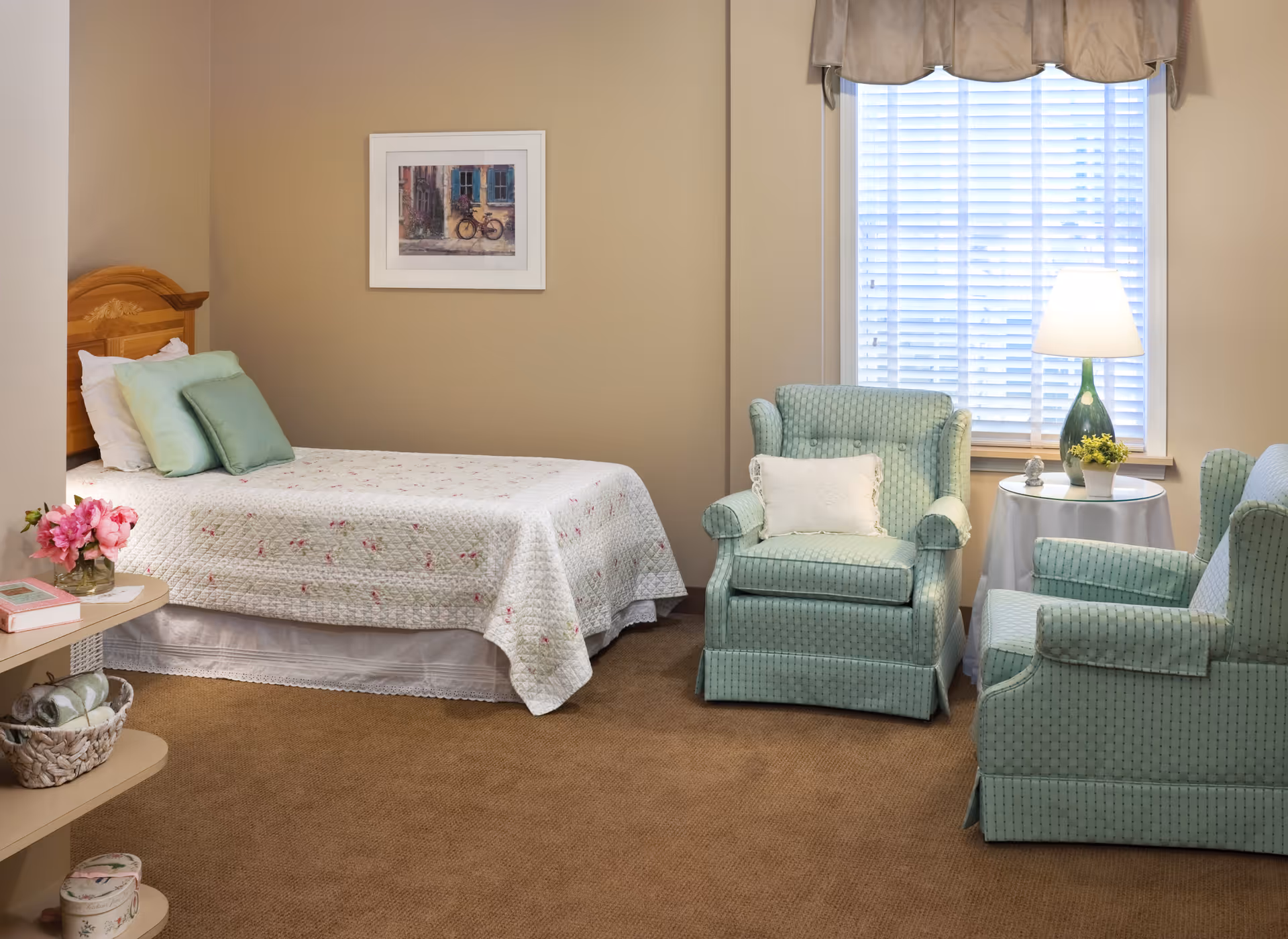 A cozy bedroom with a single bed covered in a floral quilt and green pillows. Next to the bed are two light green upholstered armchairs with a small round table between them, holding a lamp and a small plant. A window with white blinds and a beige valance is behind the chairs. A framed picture of a bicycle hangs on the beige wall above the bed. A small shelf with flowers and books is visible on the left side.
