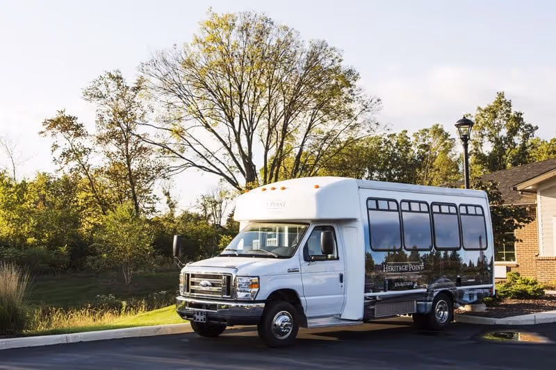 A white Heritage Point shuttle bus parked outside a senior living building with trees and landscaping in the background.