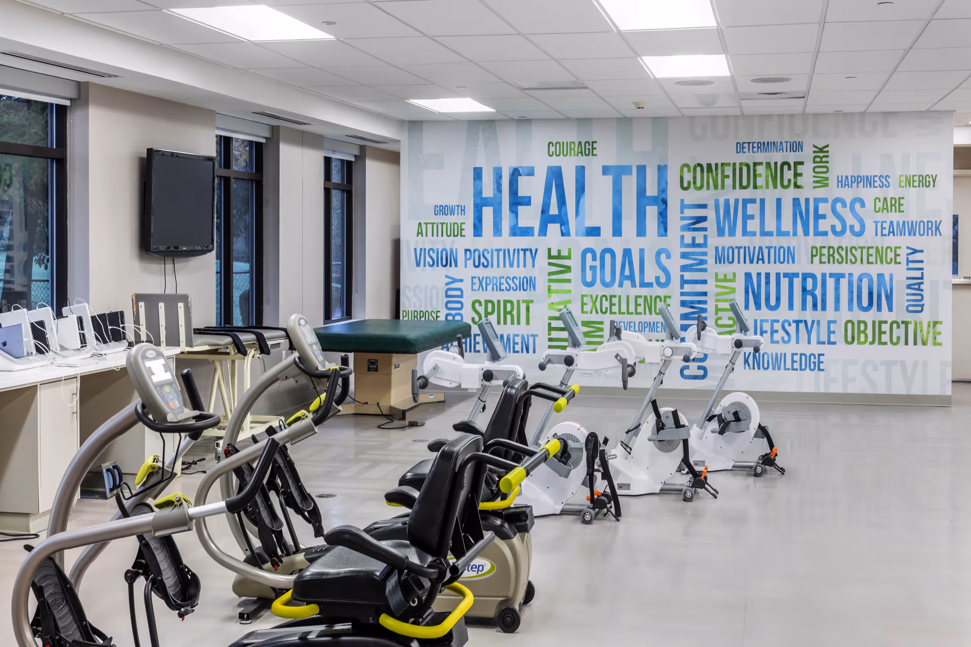 A fitness room with exercise bikes and other workout equipment. The back wall features a large mural with motivational words related to health, wellness, nutrition, and goals in blue and green text. There are windows along one side of the room and a TV mounted on the wall.