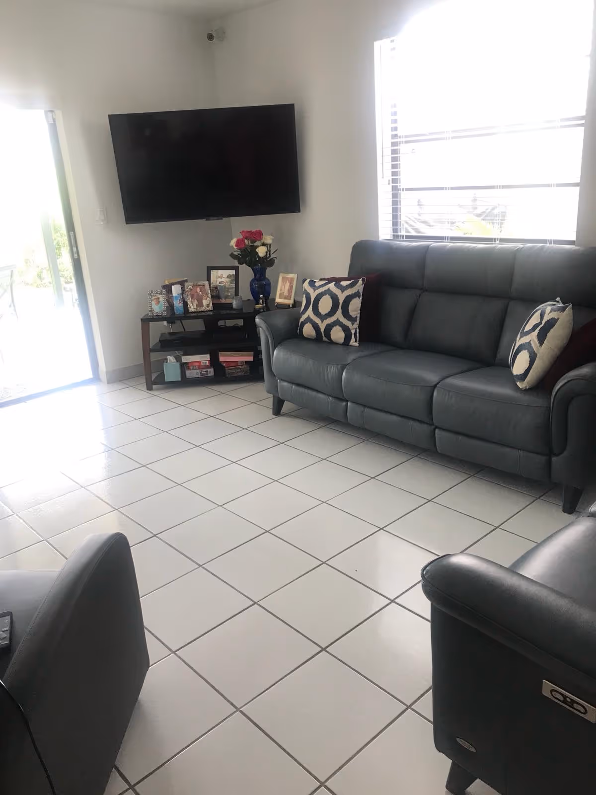 Living room with gray leather sofa and armchairs, a wall-mounted TV, a small media console with framed photos and flowers, and a tiled floor.