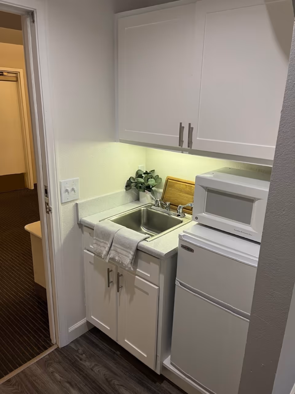 Small kitchenette area with a stainless steel sink, white cabinets above and below the counter, a white microwave on top of a white mini refrigerator, a small plant, and a wooden cutting board. A white towel hangs over the sink edge. The floor is dark wood, and a doorway leads to a carpeted hallway.