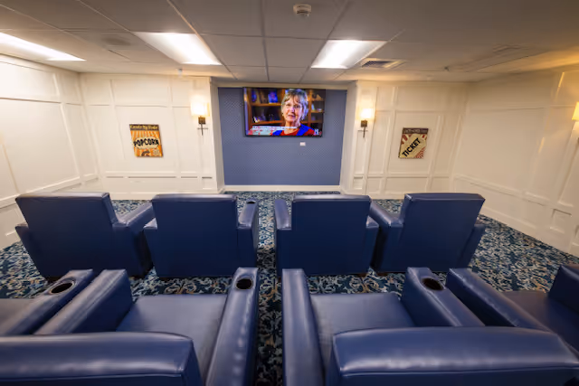 Small screening room with rows of blue leather recliners facing a wall-mounted TV showing a person on screen.