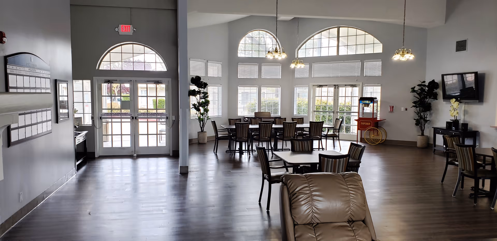 A spacious, well-lit common area with large windows and glass doors letting in natural light. The room features several tables with chairs arranged for dining or socializing, a popcorn machine in the corner, potted plants, a wall-mounted TV, and a comfortable recliner chair in the foreground. The floor is wooden, and the walls are painted light gray.