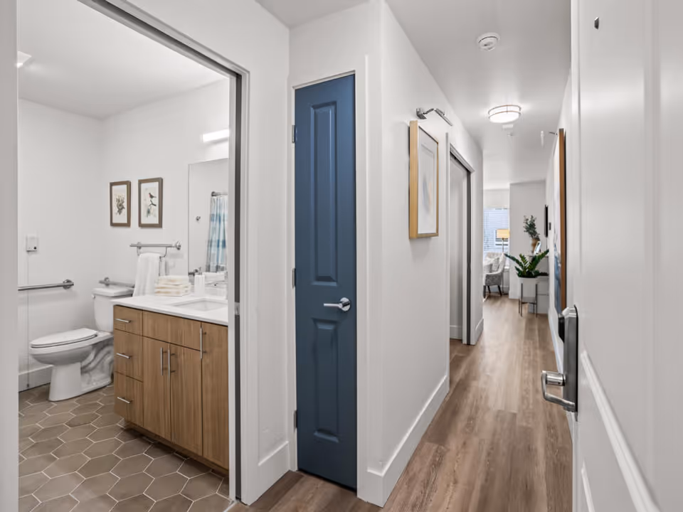 View down a hallway with light wood flooring and white walls. On the left, there is an open door leading to a bathroom with a toilet, wooden vanity with a white countertop, and framed bird artwork on the wall. A blue door is closed in the hallway. At the end of the hallway, a living area with a chair, lamp, and plants is visible.