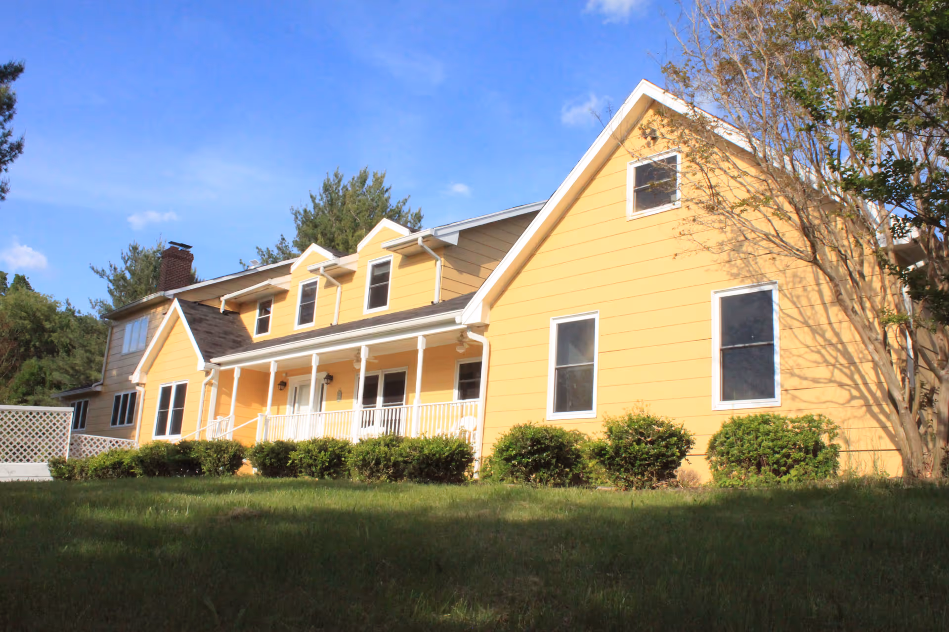 Exterior view of a large yellow two-story house with white trim, a porch with white railings, several windows, and bushes in front. The sky is clear and blue with some trees around the house.