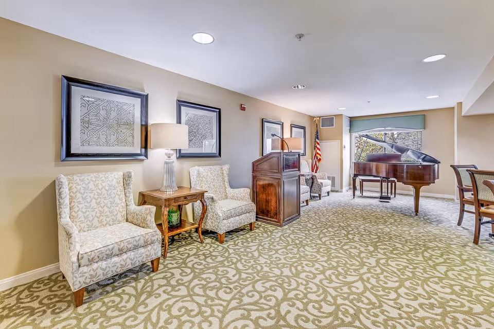 A bright and spacious common area in a senior living facility featuring patterned carpet, two upholstered armchairs with a wooden side table and lamp between them, framed artwork on the wall, a wooden podium, an American flag, a grand piano near a large window, and a dining table with chairs on the right side.