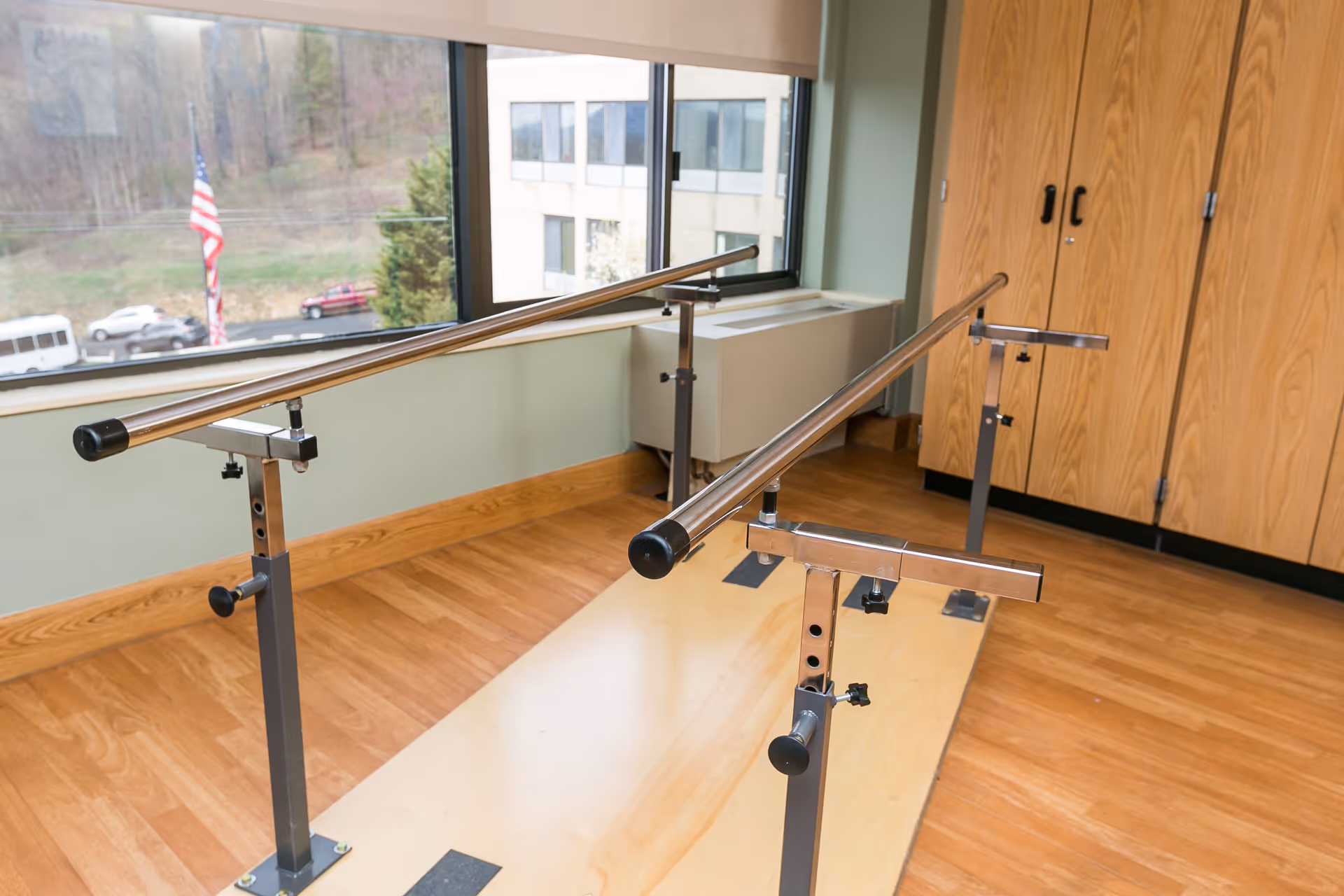 Adjustable metal parallel bars on a wooden floor in a rehabilitation room with a large window and built-in cabinets.