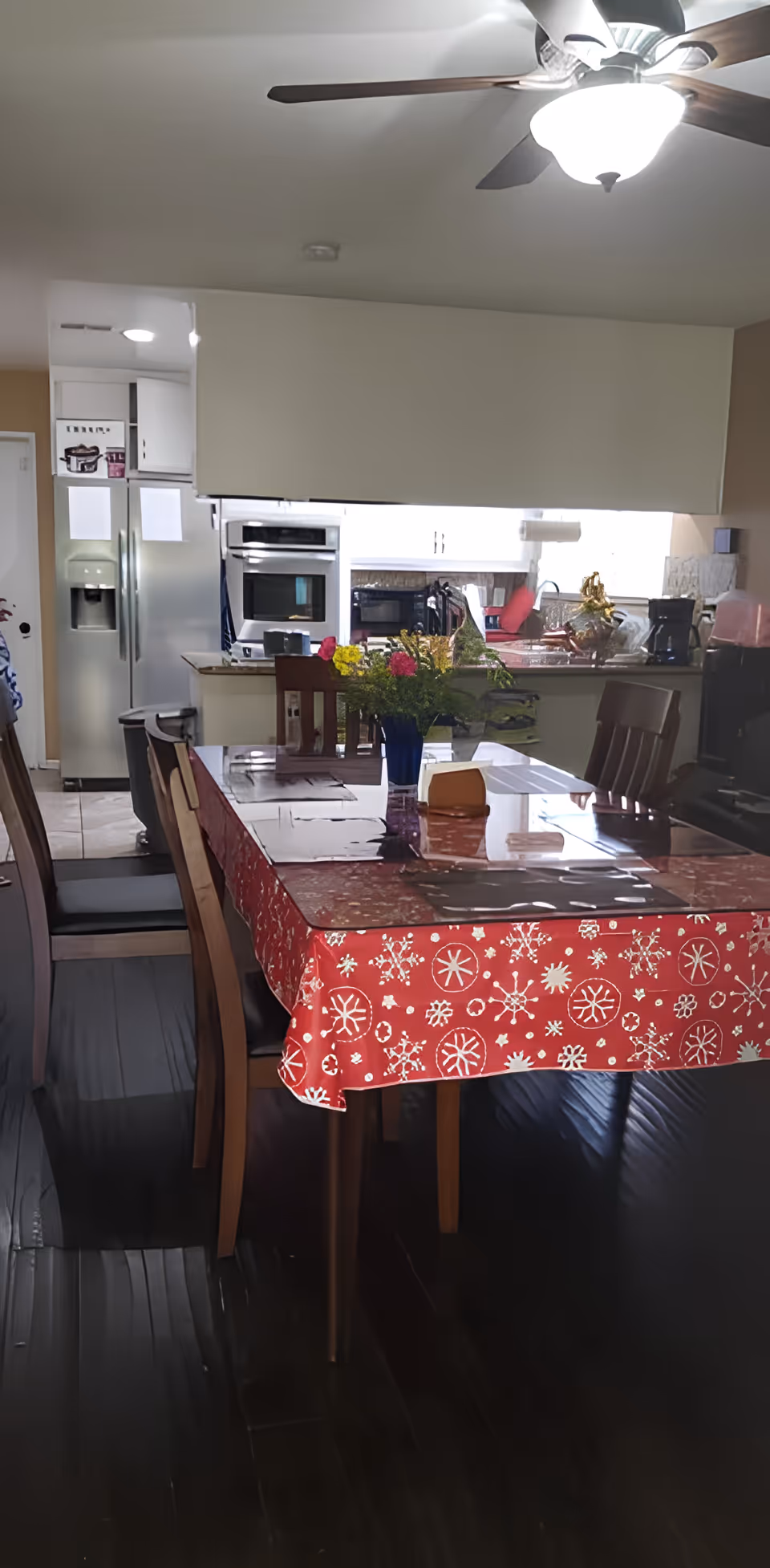 Interior view of a dining area with a wooden table covered by a red tablecloth with white snowflake patterns. The table has a glass top and a blue vase with colorful flowers in the center. Surrounding the table are wooden chairs with dark cushions. In the background, there is a kitchen area with a stainless steel refrigerator, built-in oven, microwave, and various kitchen appliances. A ceiling fan with a light is visible above.