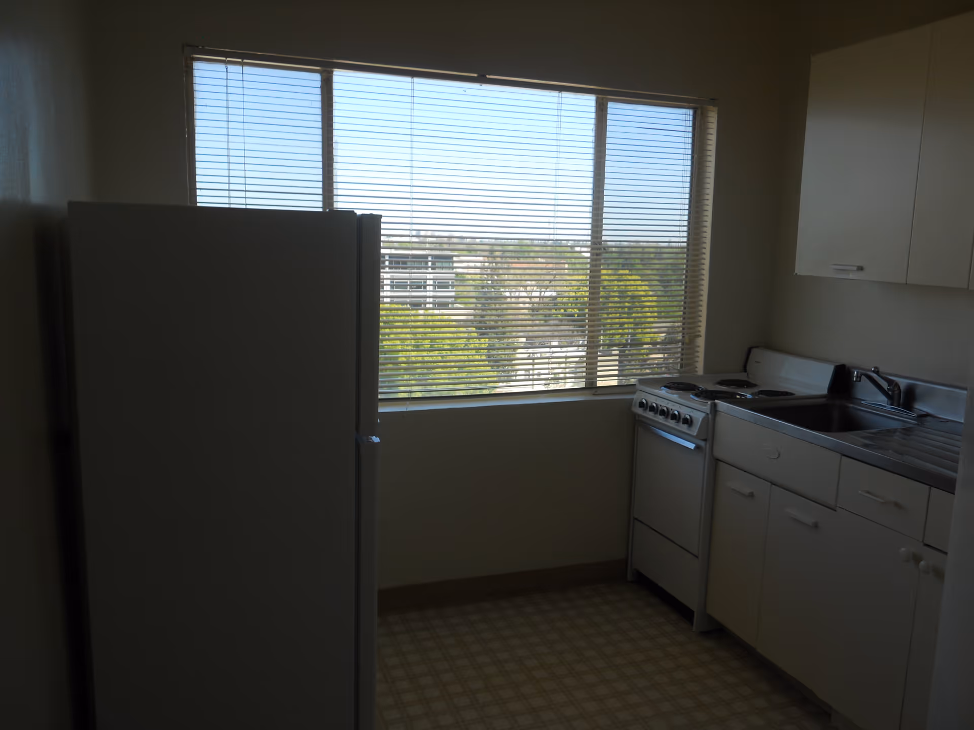 Small apartment kitchen with a refrigerator, stove, sink, cabinets and a large window with blinds.