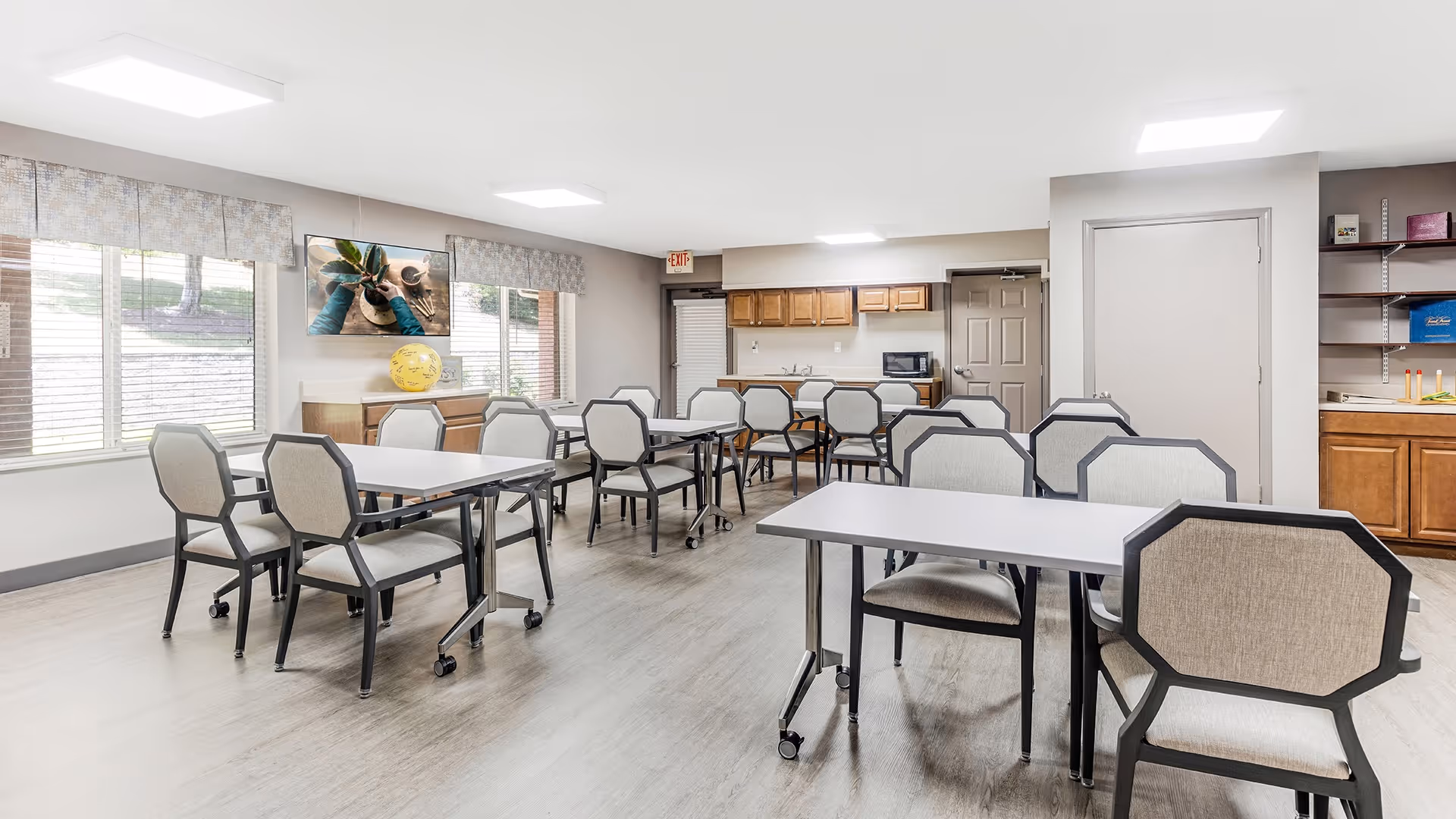 A bright and clean dining room with several tables and chairs arranged neatly. The room has large windows with blinds and valances, a wall-mounted TV, and a sideboard with decorative items. The back of the room features a kitchenette area with wooden cabinets, a microwave, and a closed door.