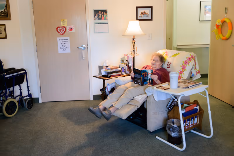 An elderly woman reclining in a beige armchair in a cozy room, reading a book titled 'Buckskin Trail'. The room has a side table with a lamp, books, and personal items. A walker is visible near a closed door with reminders and decorations. A small white table holds a cup and newspapers, with a basket underneath. The walls have framed photos and a decorative wreath.