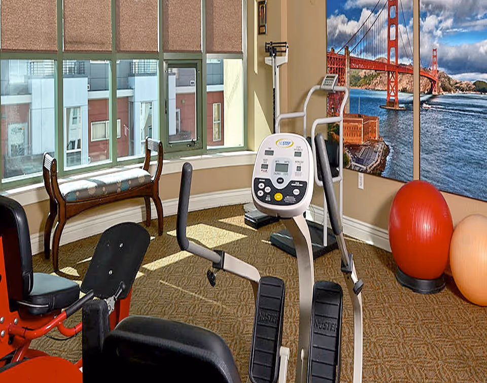 A small exercise room with various fitness equipment including a recumbent bike, an elliptical machine, and two large exercise balls. The room has large windows with beige blinds, a wooden bench, and a wall mural of the Golden Gate Bridge over water.