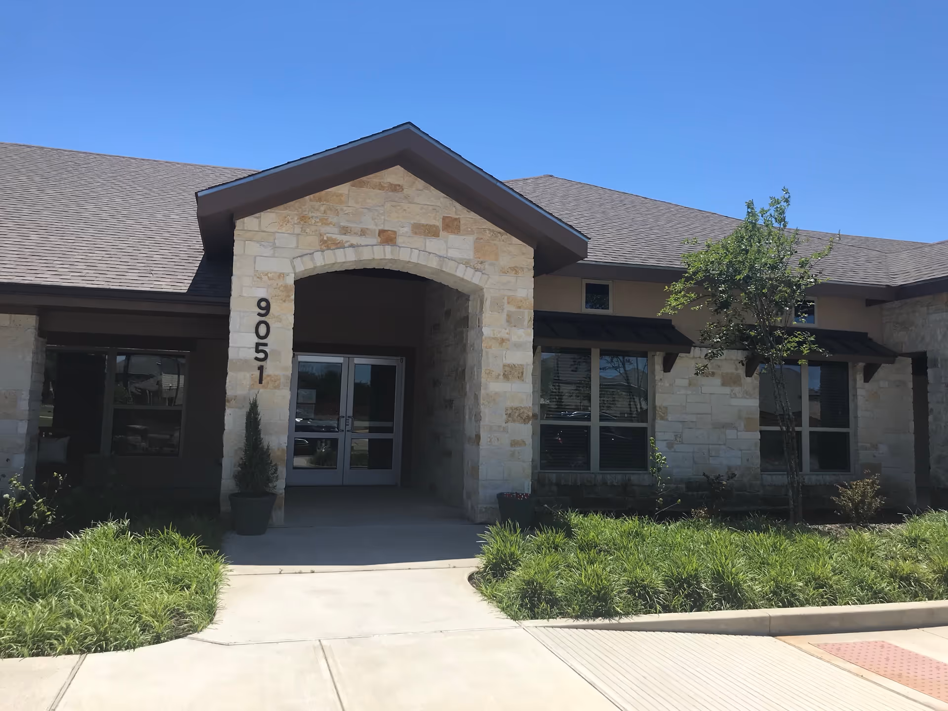 Front entrance of a single-story stone-faced building with double doors, address number 9051, and landscaping.