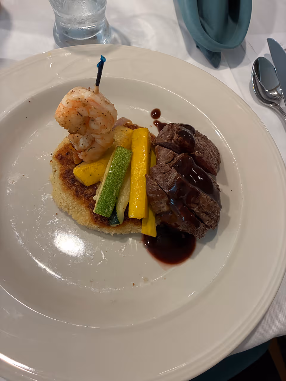 A plated meal featuring two pieces of cooked shrimp on a skewer, grilled yellow and green zucchini strips, a round piece of toasted bread or polenta, and two slices of steak topped with a dark sauce, served on a white plate with a glass of water and silverware in the background.