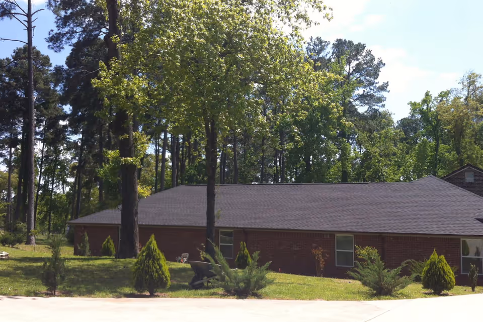Single-story brick building with a dark roof surrounded by green trees and shrubs under a clear blue sky.