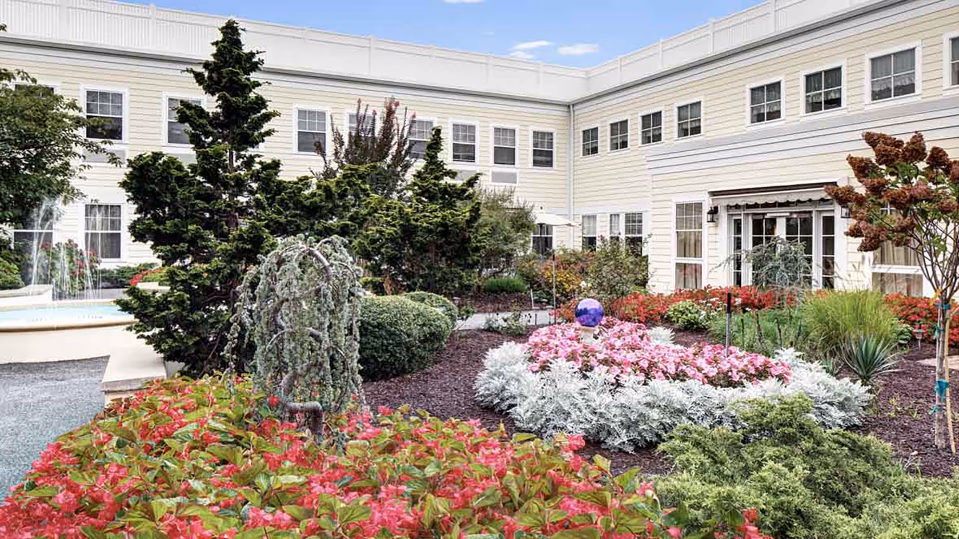 Landscaped courtyard with colorful flower beds, shrubs, and a fountain in front of a pale yellow multi-story senior living building.
