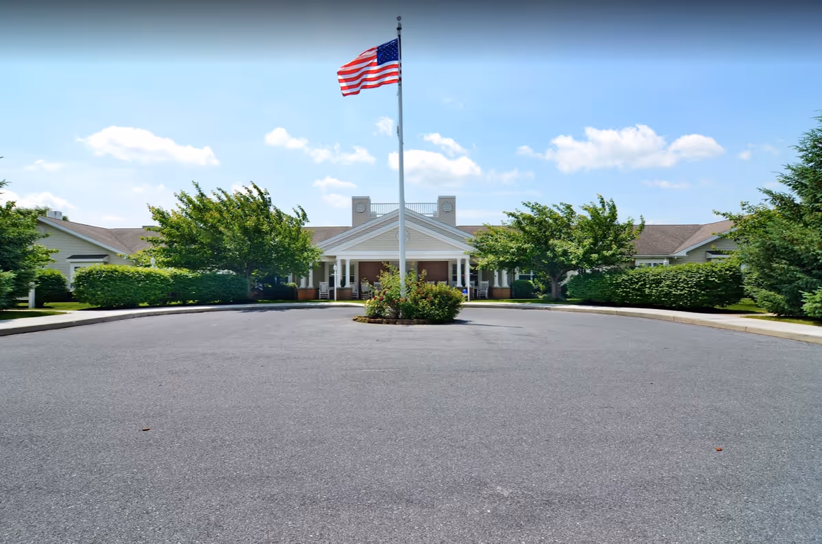 Front exterior view of Celebration Villa of Lebanon PA, showing a single-story building with a covered entrance, an American flag on a flagpole in the center, surrounded by trees and bushes under a blue sky with some clouds.