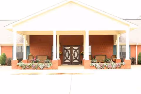 Front entrance of a brick senior living facility with benches and flowers.
