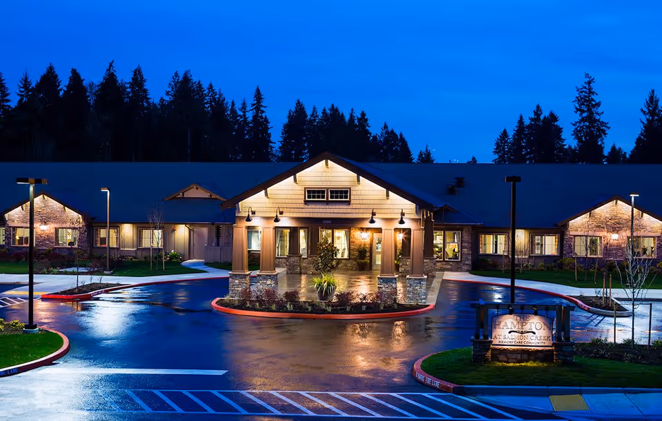 The Hampton at Salmon Creek Memory Care Community building exterior at dusk, with warm lights illuminating the entrance and surrounding windows. The driveway and parking area are wet, reflecting the lights, and tall trees are visible in the background under a deep blue sky.