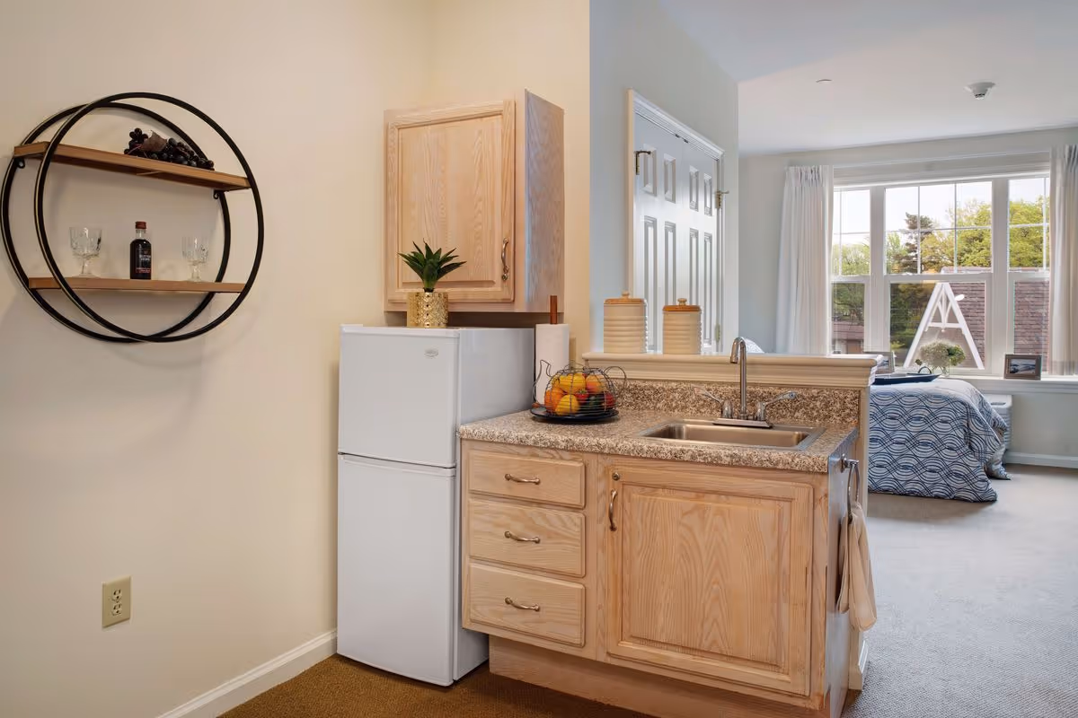 A small kitchenette area with a white mini refrigerator, wooden cabinets, and a granite countertop with a sink. On the countertop, there is a fruit basket and two canisters. To the left, a circular wall shelf holds two wine glasses and a bottle. In the background, a bedroom with a bed covered in a blue patterned bedspread is visible next to a large window with white curtains, letting in natural light.