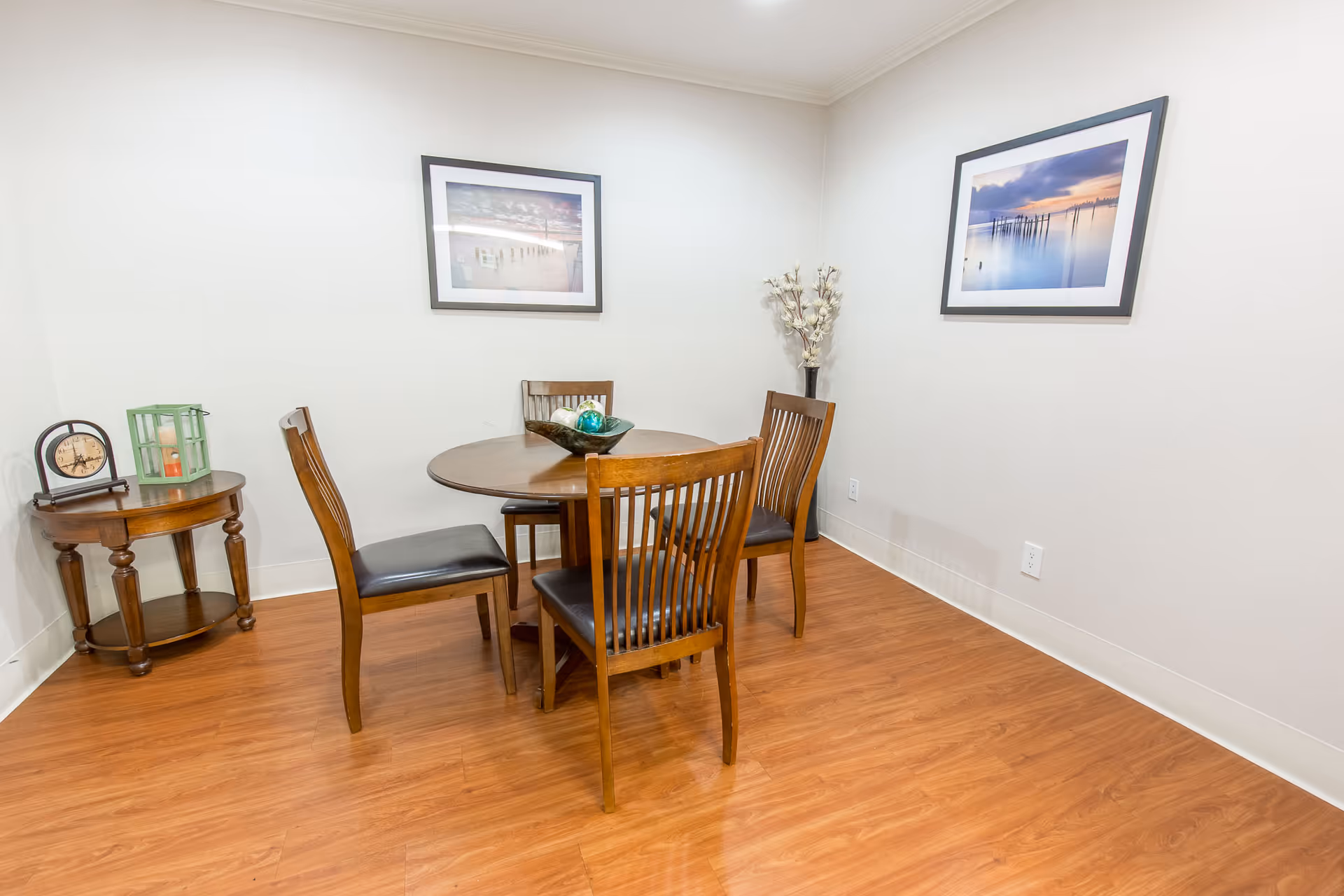A small dining area with a round wooden table surrounded by four wooden chairs with black cushions. On the table is a decorative bowl with ornaments. To the left, there is a small round wooden side table with a clock and a green lantern. The walls are white and decorated with two framed landscape photographs. The floor is a light wood laminate.