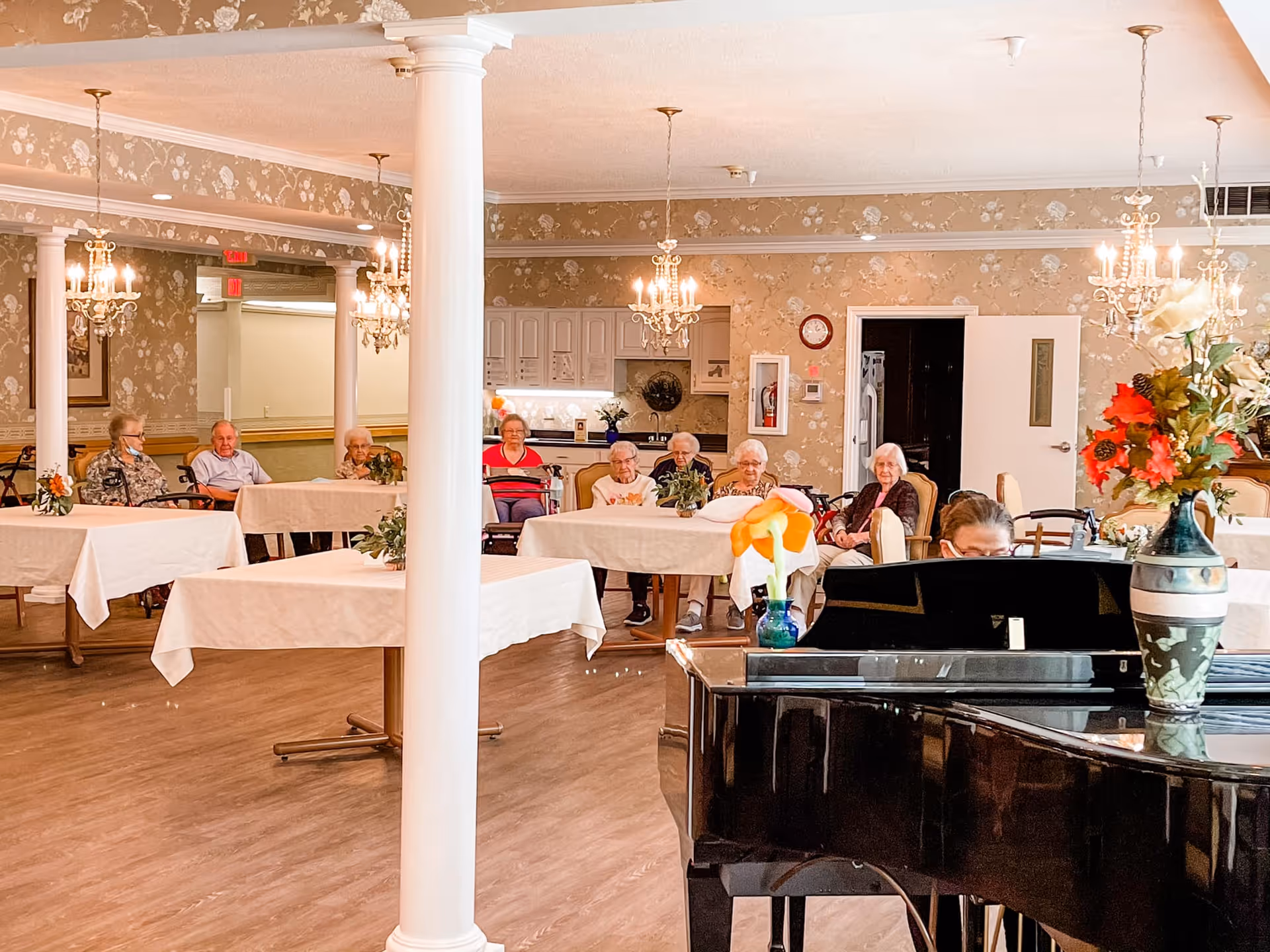 A group of elderly people sitting at tables covered with white tablecloths in a decorated room with floral wallpaper and chandeliers. A black grand piano with flower vases on top is in the foreground, and a person is playing the piano. The room has wooden flooring and white columns.