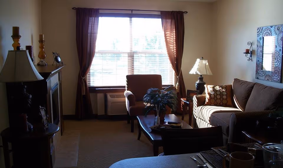 A cozy living room with a large window covered by brown curtains letting in natural light. The room features a brown sofa with patterned cushions, an armchair, a wooden coffee table with a plant on it, a side table with a lamp, and a fireplace with decorative items on the mantel. A mirror and wall sconces are mounted on the wall above the sofa.
