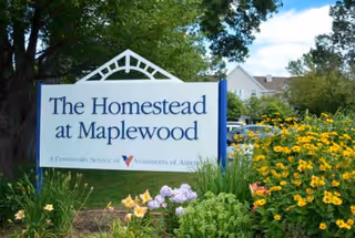 Outdoor sign for The Homestead at Maplewood surrounded by colorful flowers and greenery with trees and a building in the background under a partly cloudy sky.
