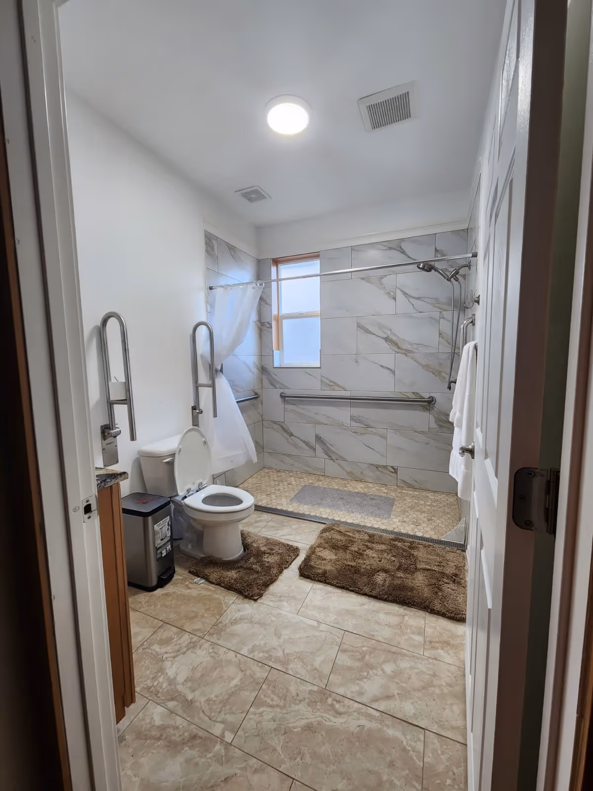 A clean and spacious bathroom with beige tiled floor and walls. It features a toilet with the lid open, two brown bath mats on the floor, a trash bin, and a walk-in shower area with a curtain and grab bars. The shower walls have large marble-patterned tiles and a window letting in natural light. The ceiling has a round light fixture and ventilation vents.