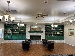 A spacious living room with a fireplace centered on a wall with green built-in shelves on either side. The shelves contain decorative items and books. There are four black armchairs arranged around the fireplace. The ceiling has multiple light fixtures including chandeliers and a ceiling fan. The floor is wooden, and there is a framed picture on the right wall.