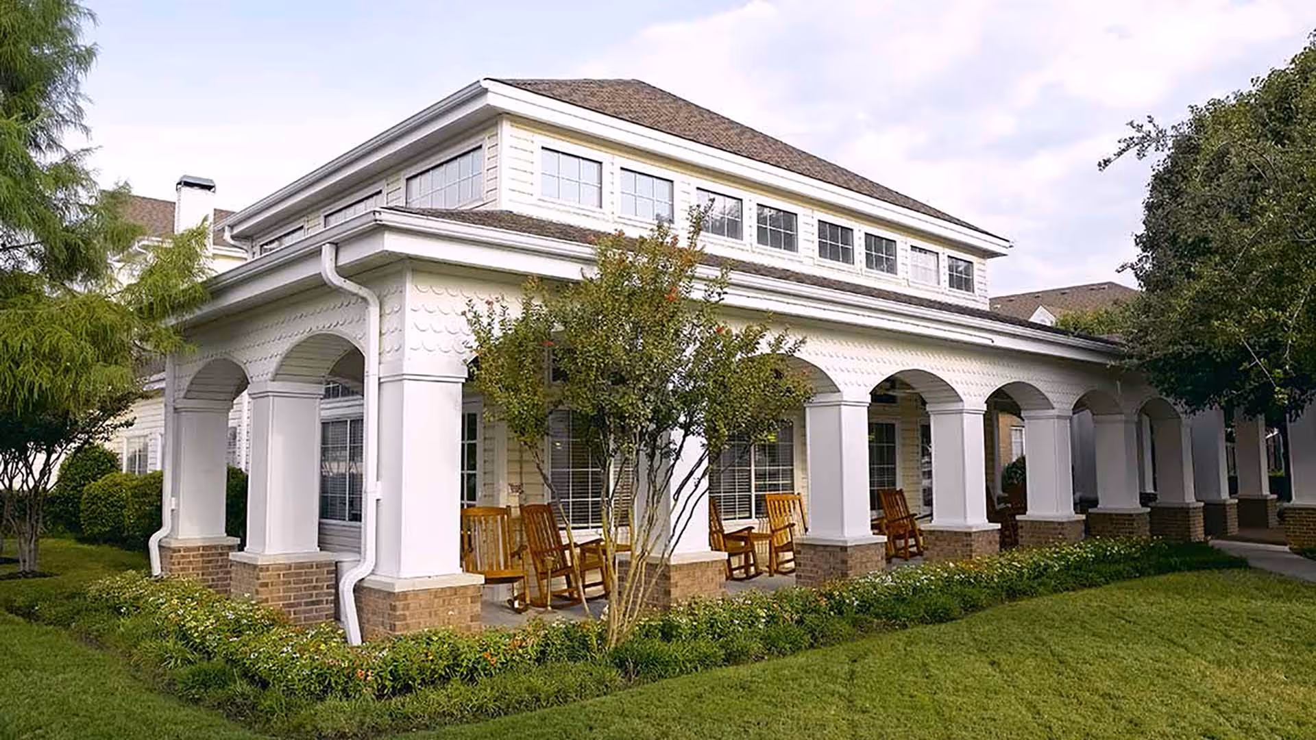 Exterior view of a senior living facility building with white columns and arches, a covered porch with wooden rocking chairs, surrounded by green grass, bushes, and trees under a partly cloudy sky.