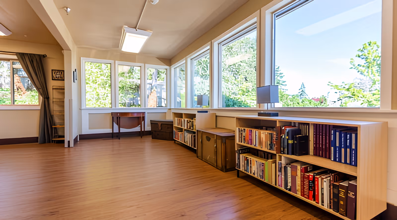 Bright common room with large windows, low bookshelves filled with books, and a wood floor.
