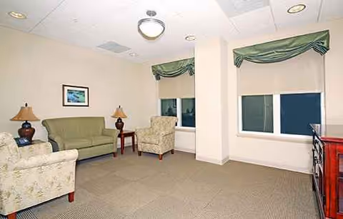 Lounge area with sofa, chairs, lamps, and windows in a senior care facility.