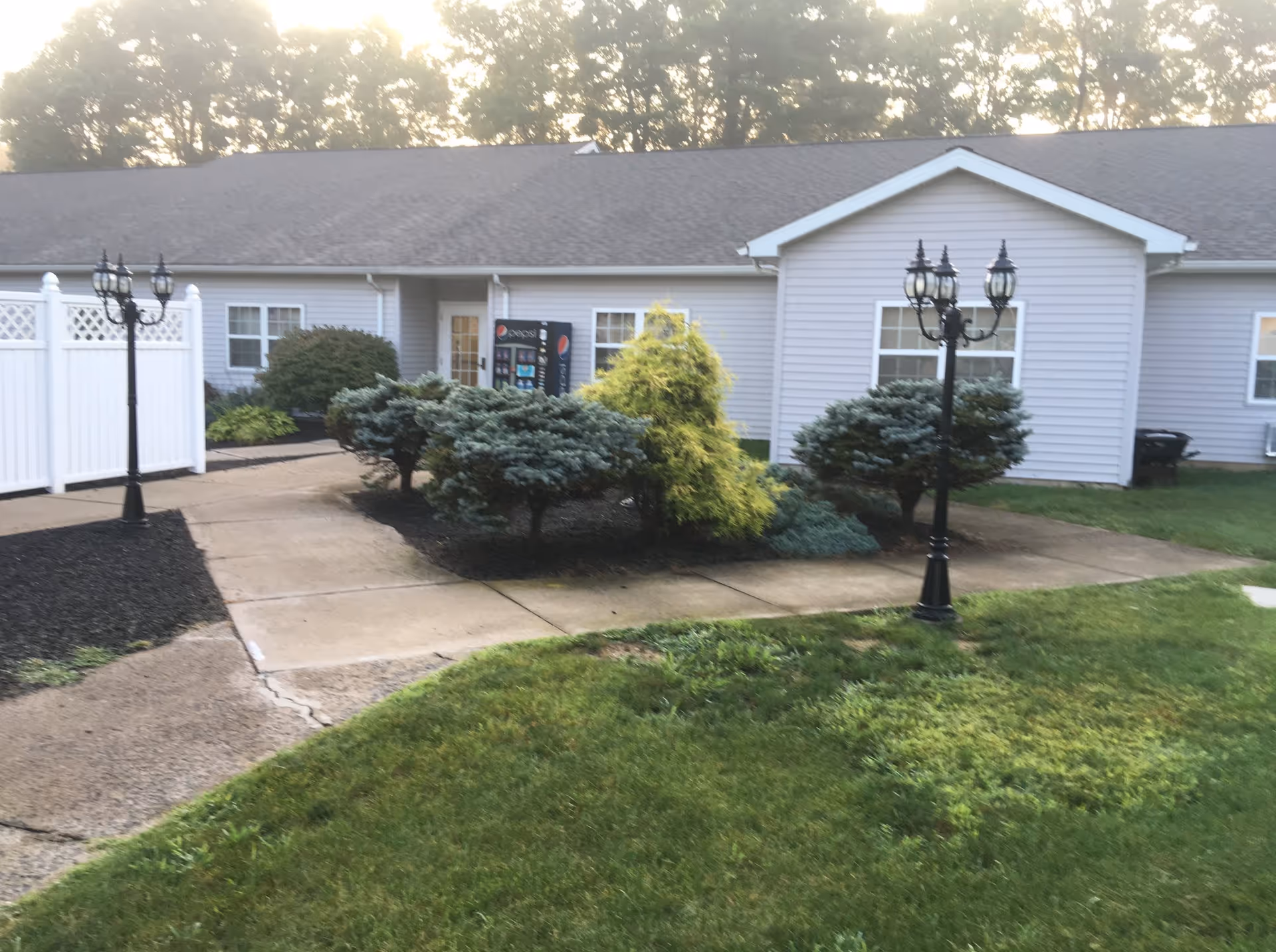 Front entrance of a single-story senior living building with a walkway, lamp posts, shrubs, and a vending machine visible by the door.
