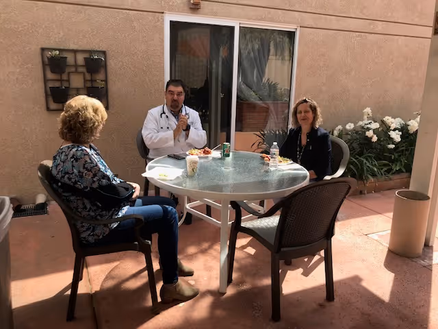 Three people sitting around a round glass table outdoors, eating and talking. There are plants and flowers along the wall behind them, and a sliding glass door in the background.