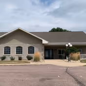 Front exterior view of a single-story senior living facility building with beige siding and stone accents, a covered entrance, and a paved driveway in front under a cloudy sky.