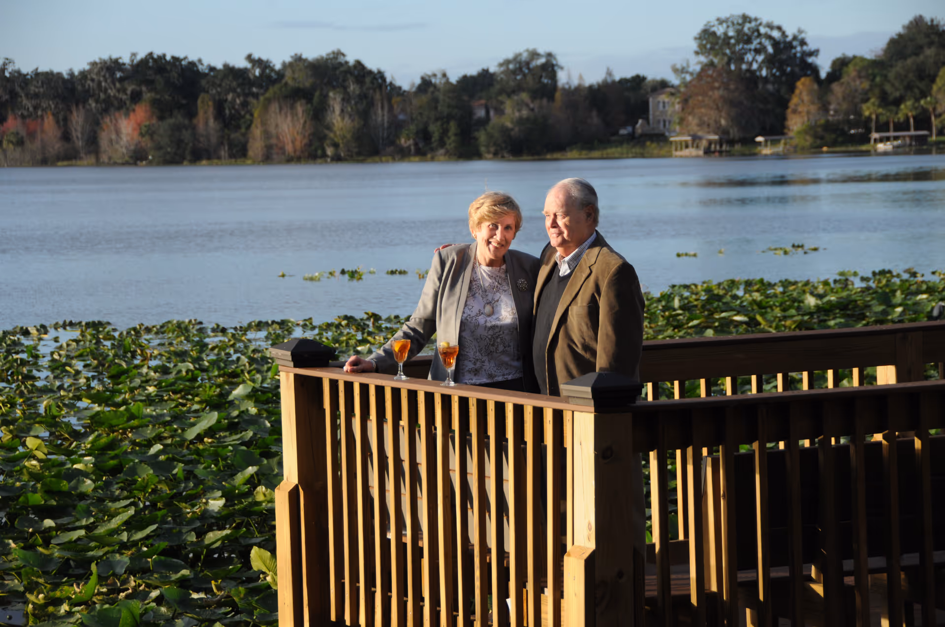 An elderly couple standing on a wooden deck overlooking a lake with lily pads and trees in the background. They have two glasses of an orange-colored drink on the railing beside them.